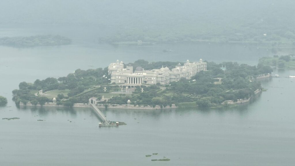 Panoramic view of Raffles Udaipur's main island building from a nearby mountain, with a historic 100-year-old Shiva Temple visible in the foreground