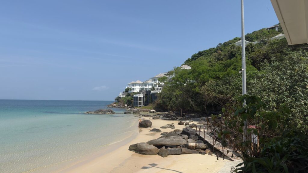 Premier Village Phu Quoc Resort private beach in the afternoon – turquoise sea, rocky shoreline, white villas nestled in lush greenery on the hillside, Phu Quoc Island Vietnam